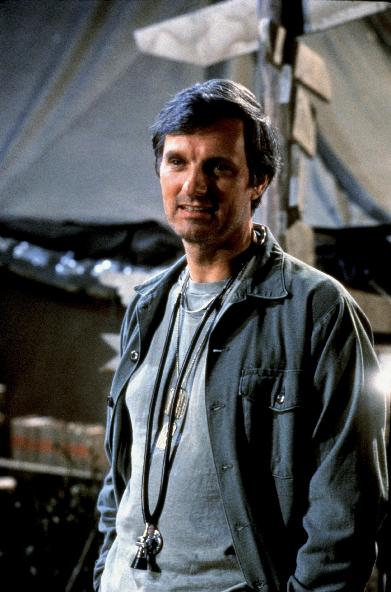 Mężczyzna w średnim wieku w lekkiej kurtce i koszulce, na szyi ma nieco dłuższy naszyjnik z metalowymi zawieszkami, w tle namiot oraz rozmazane drewniane znaki.