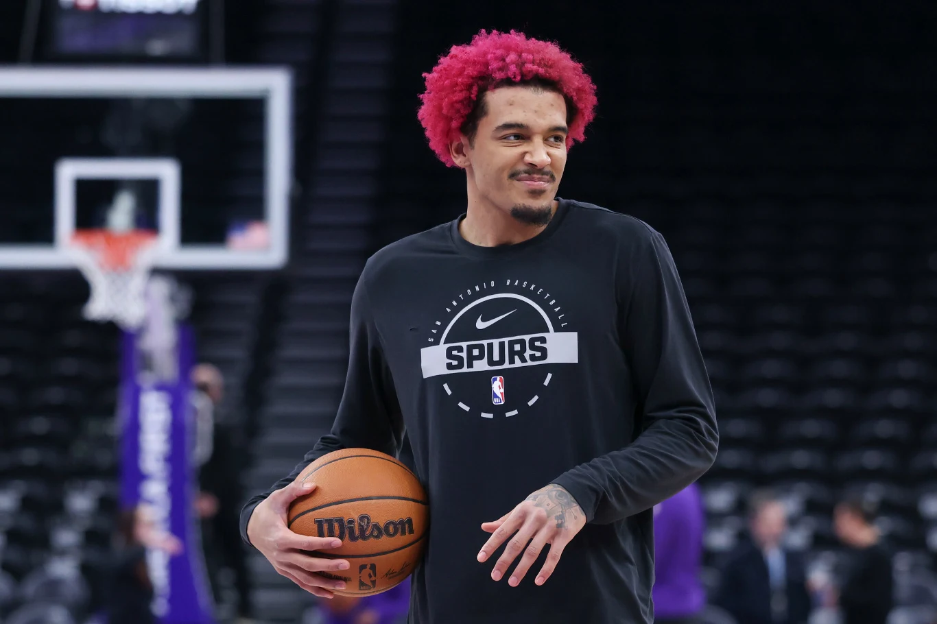 Młody mężczyzna z czerwonymi włosami, ubrany w czarną koszulkę klubu Spurs, trzyma piłkę do koszykówki na hali sportowej. W tle rozmazane sylwetki osób i puste trybuny.