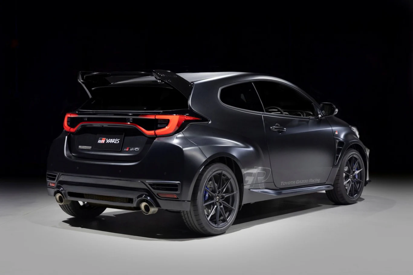 Czarny, sportowy samochód z masywnym tylnym spojlerem, podwójnym układem wydechowym i felgami z wieloma ramionami, ustawiony na ciemnym tle, podkreślającym jego agresywną sylwetkę.