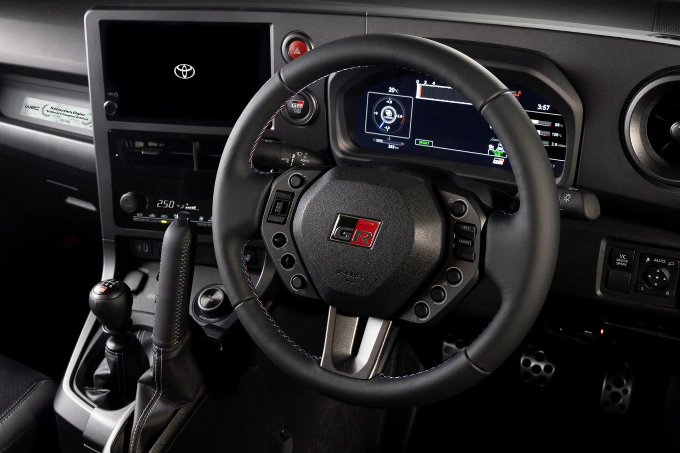 Nowoczesna deska rozdzielcza samochodu z logo Toyota GR, widoczną kierownicą sportową z przyciskami, zaawansowanym cyfrowym wyświetlaczem, manualną skrzynią biegów, pedałami oraz nowoczesnym panelem sterowania.