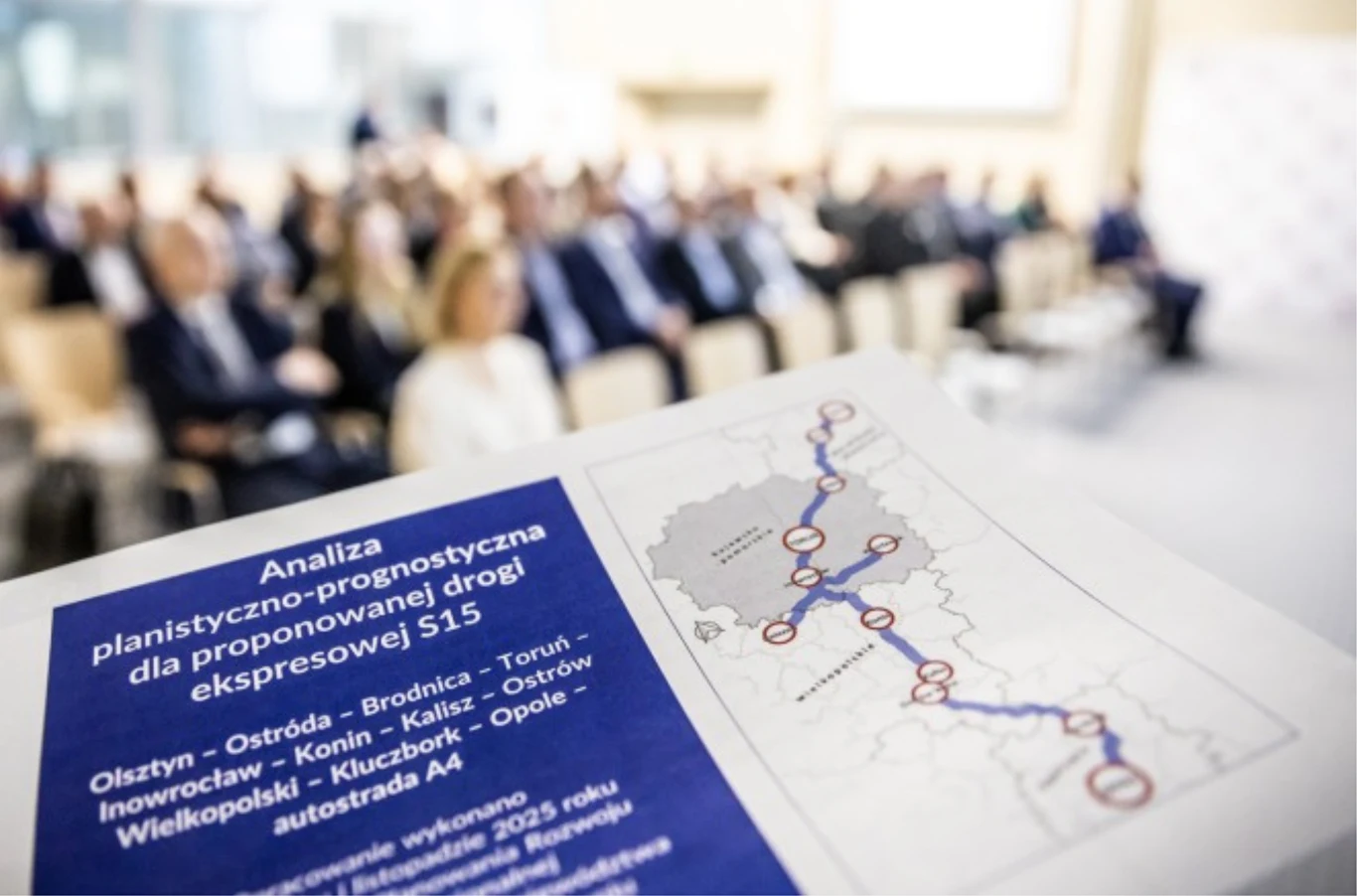 Widok sali konferencyjnej z uczestnikami siedzącymi w rzędach oraz dokumentem z mapą przedstawiającą analizę planistyczno-prognostyczną dotyczącą projektowanej drogi ekspresowej S15, umieszczonym na pierwszym planie.