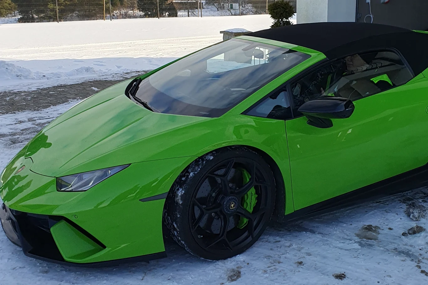 Jaskrawozielony sportowy samochód marki Lamborghini z czarnym dachem stoi na ośnieżonej nawierzchni, widoczne czarne felgi oraz detale aerodynamiczne, w tle zimowy krajobraz.