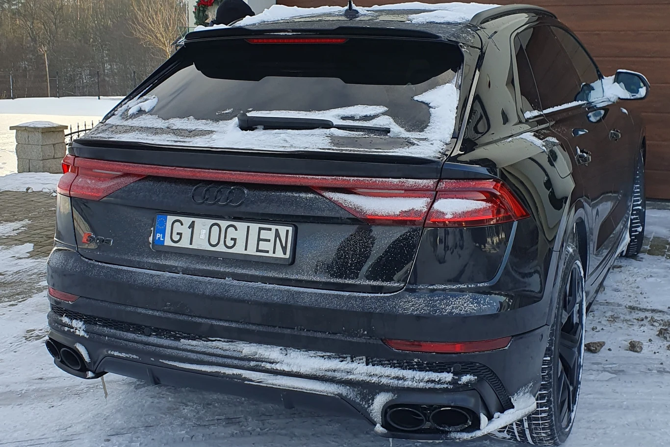 Czarny samochód marki Audi SUV zaparkowany na ośnieżonej drodze przy drewnianym budynku, lekko przysypany śniegiem, z widoczną charakterystyczną tablicą rejestracyjną, zimowy krajobraz w tle.