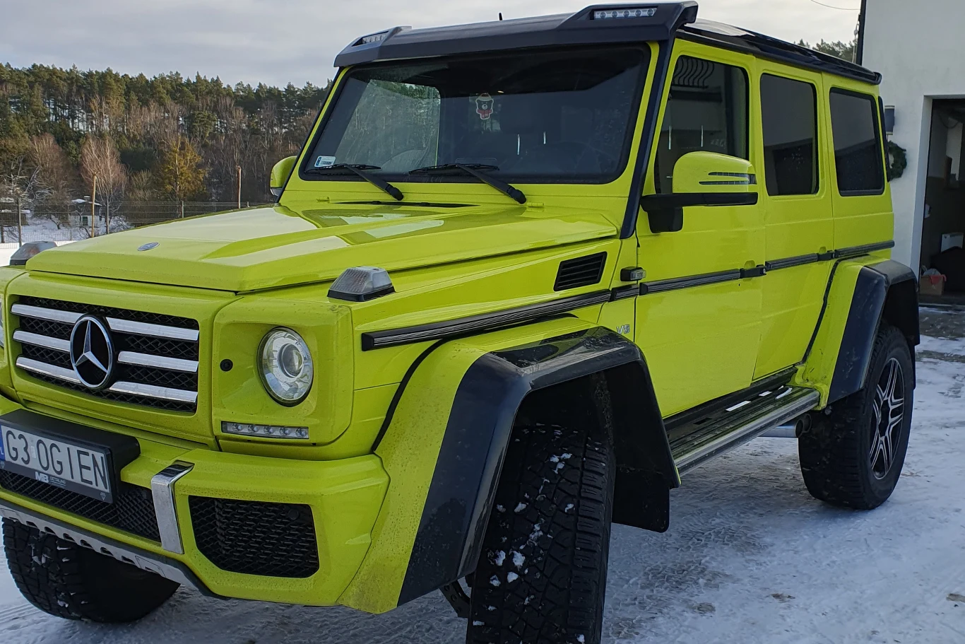 Jaskrawy, żółtozielony Mercedes klasy G na terenowych oponach stoi zaparkowany na śniegu, w tle las oraz fragment budynku.
