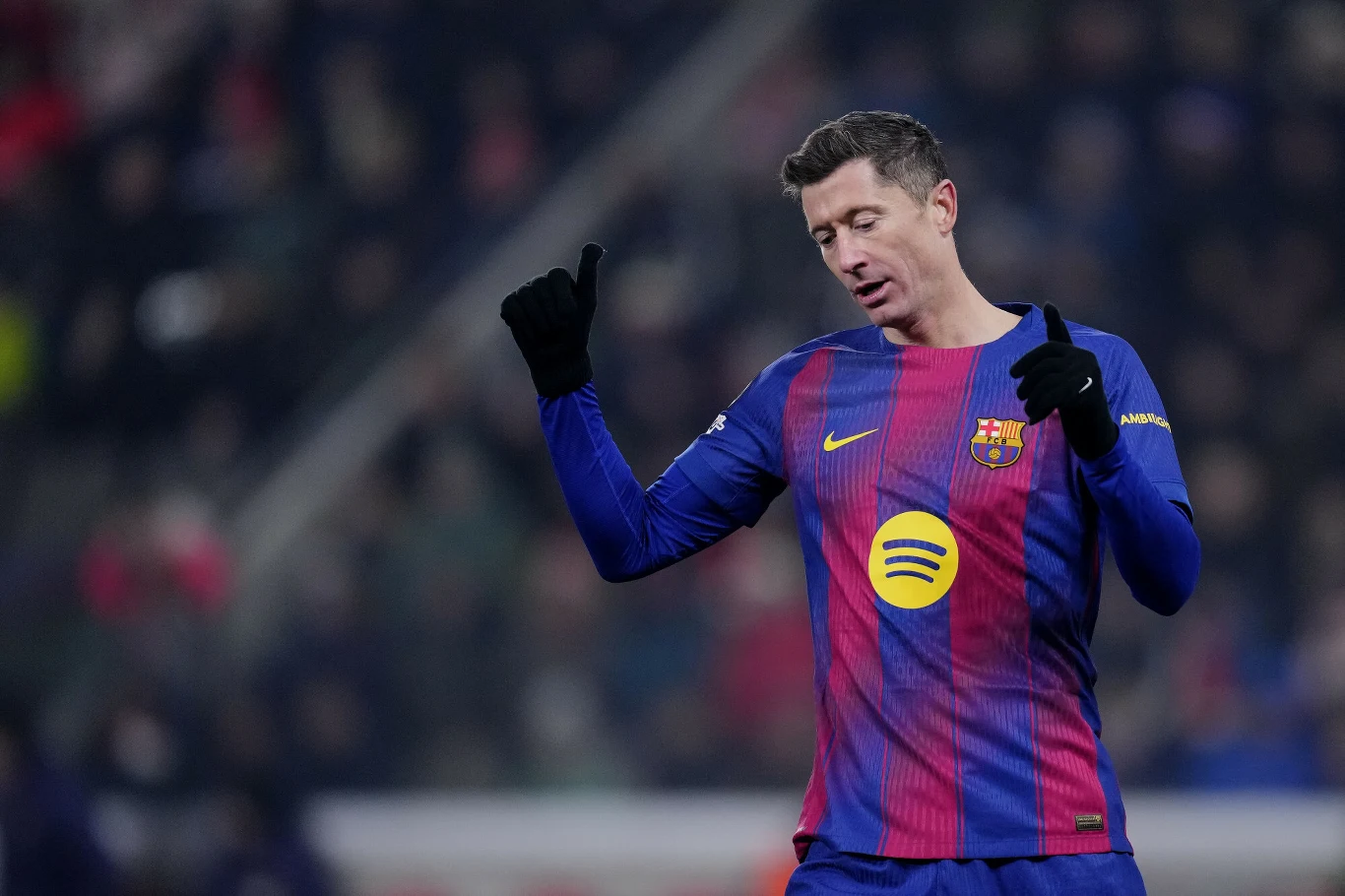 Piłkarz w niebiesko-czerwonej koszulce FC Barcelony z logo sponsora na piersi, w czarnych rękawiczkach, wykonuje gesty rękami podczas meczu na stadionie, w tle rozmyta publiczność.