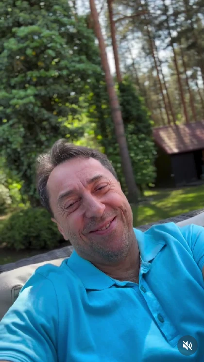 Uśmiechnięty mężczyzna w jasnoniebieskiej koszulce z krótkim rękawem odpoczywa na zewnątrz, w tle drzewa iglaste i domek ogrodowy, jasne światło słoneczne podkreśla pogodny nastrój sceny.