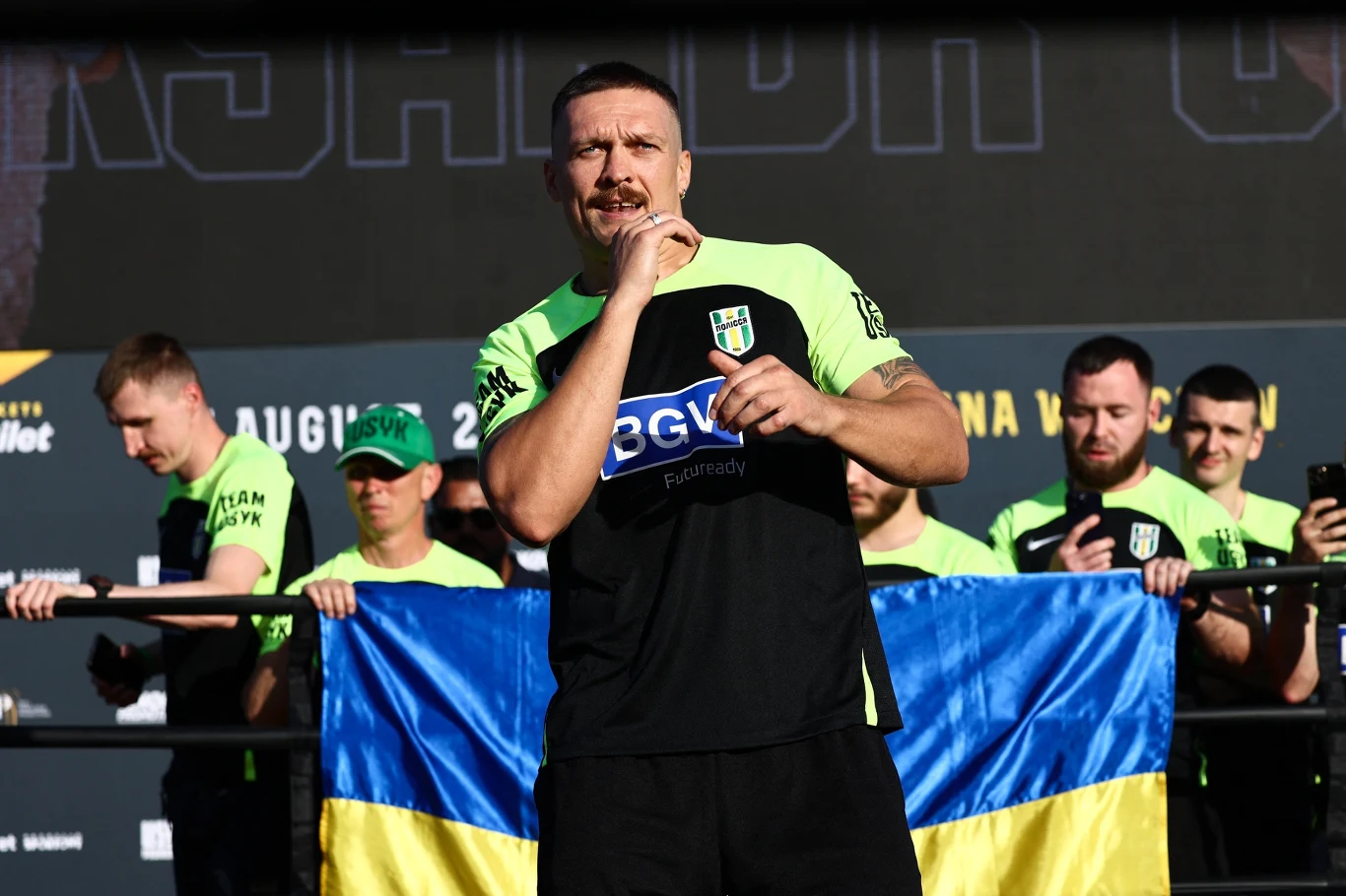 Ołeksandr Usyk Mężczyzna w sportowym stroju stoi na scenie, trzymając mikrofon przy ustach, z tyłu grupa ludzi w zielonych koszulkach oraz trzymana flaga Ukrainy.