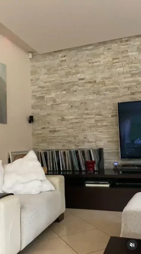 Nowoczesny salon z jasnymi kanapami, ścianą wykończoną dekoracyjnym kamieniem i telewizorem po prawej stronie. Na niskiej szafce ustawione są płyty winylowe i czerwony wazon.