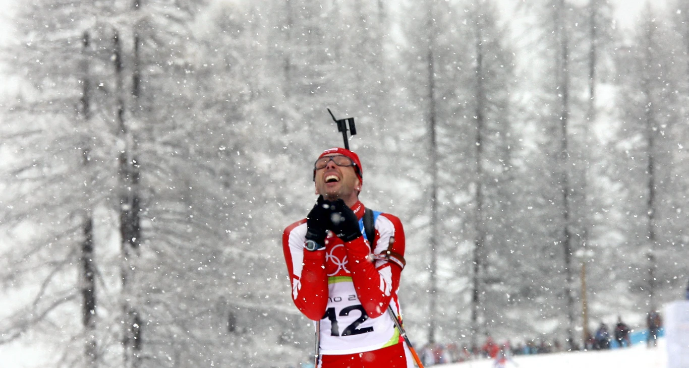 Zawodnik biathlonowy w czerwono-białym stroju z numerem startowym na piersi stoi na śniegu w otoczeniu padającego śniegu, unosząc głowę i zaciśnięte dłonie w geście radości lub ulgi, w tle ośnieżone drzewa.