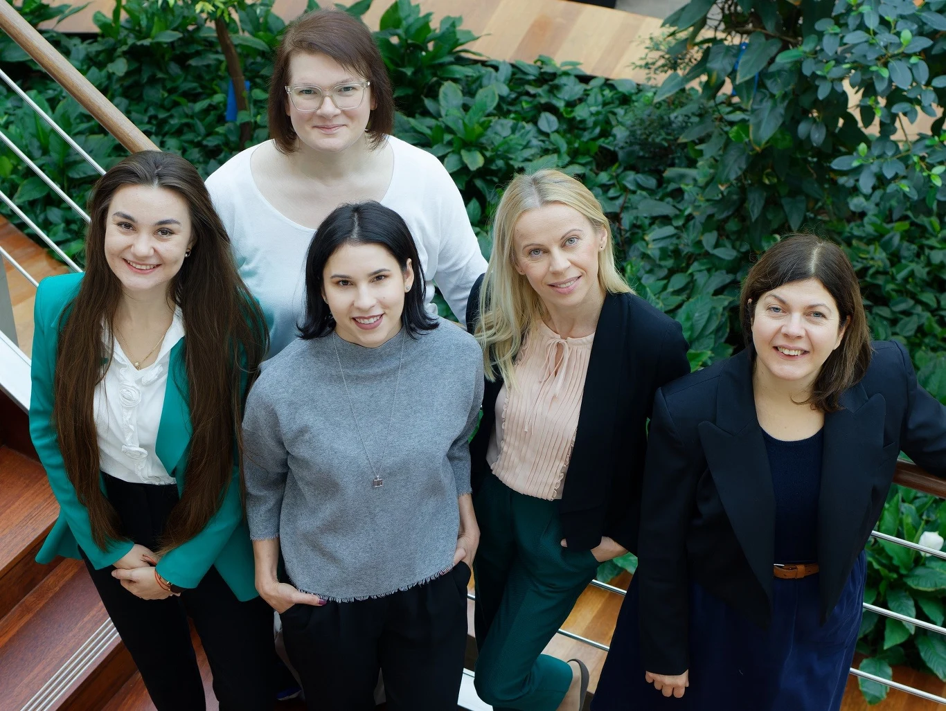 Na fotografii stoją od lewej współtwórczynie technologii: mgr Aleksandra Krajcer, dr n med. Ewelina Grzywna (z tyłu), dr Alicja Hinz (z przodu), dr hab. Joanna Lewandowska-Łańcucka, prof. UJ, dr hab. Monika Bzowska, prof. UJ. Pięć kobiet stojących razem na schodach, ubrane elegancko i uśmiechnięte, otoczone zielenią doniczkowych roślin oraz drewnianą balustradą.