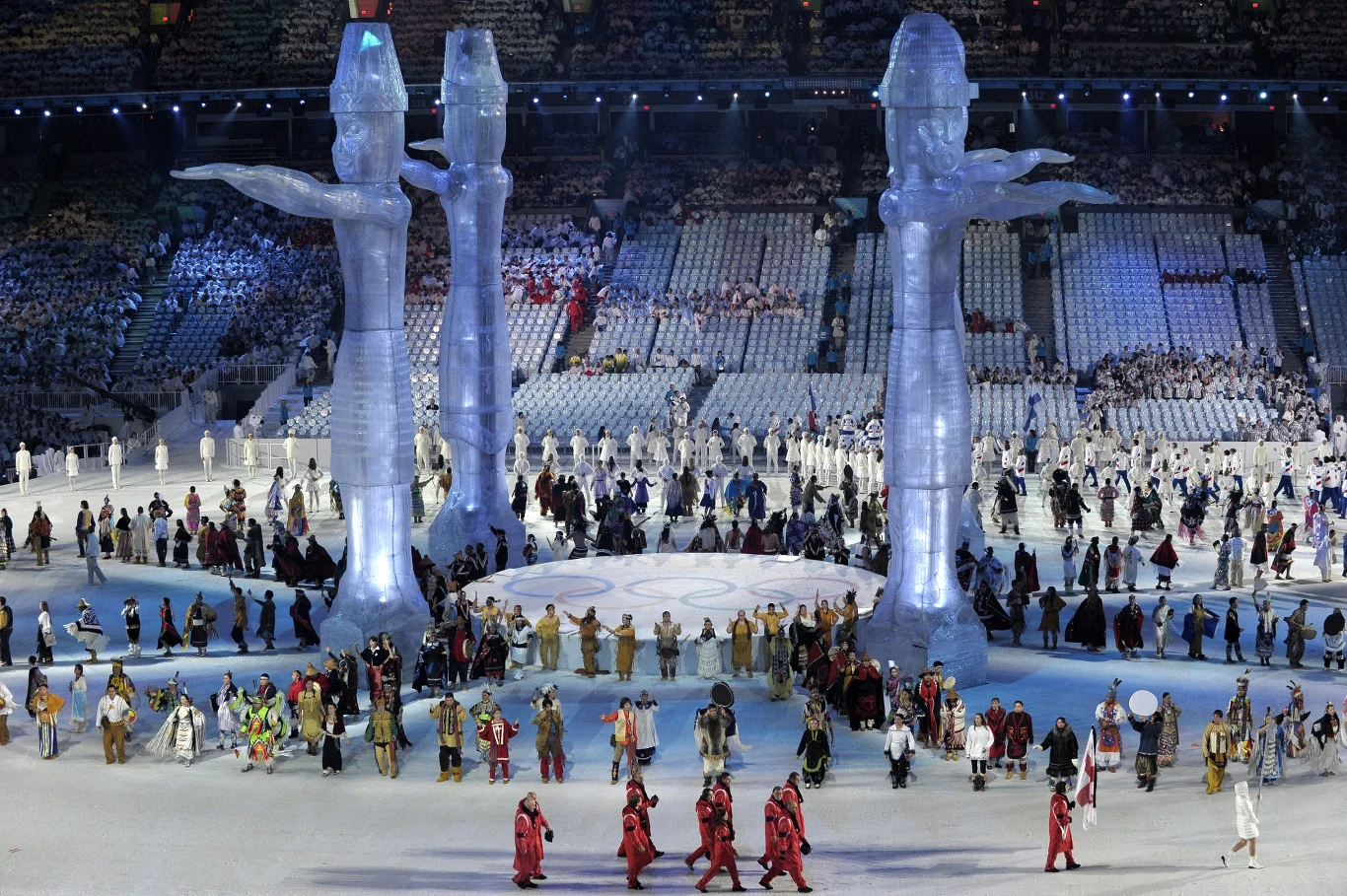 Gruzińscy sportowcy na ceremonii otwarcia igrzysk pojawili się z czarnymi opaskami Kolorowa parada ludzi ubranych w tradycyjne stroje, ustawionych wokół ogromnych przezroczystych rzeźb przedstawiających postacie z uniesionymi ramionami na stadionie, trybuny zapełnione widzami.