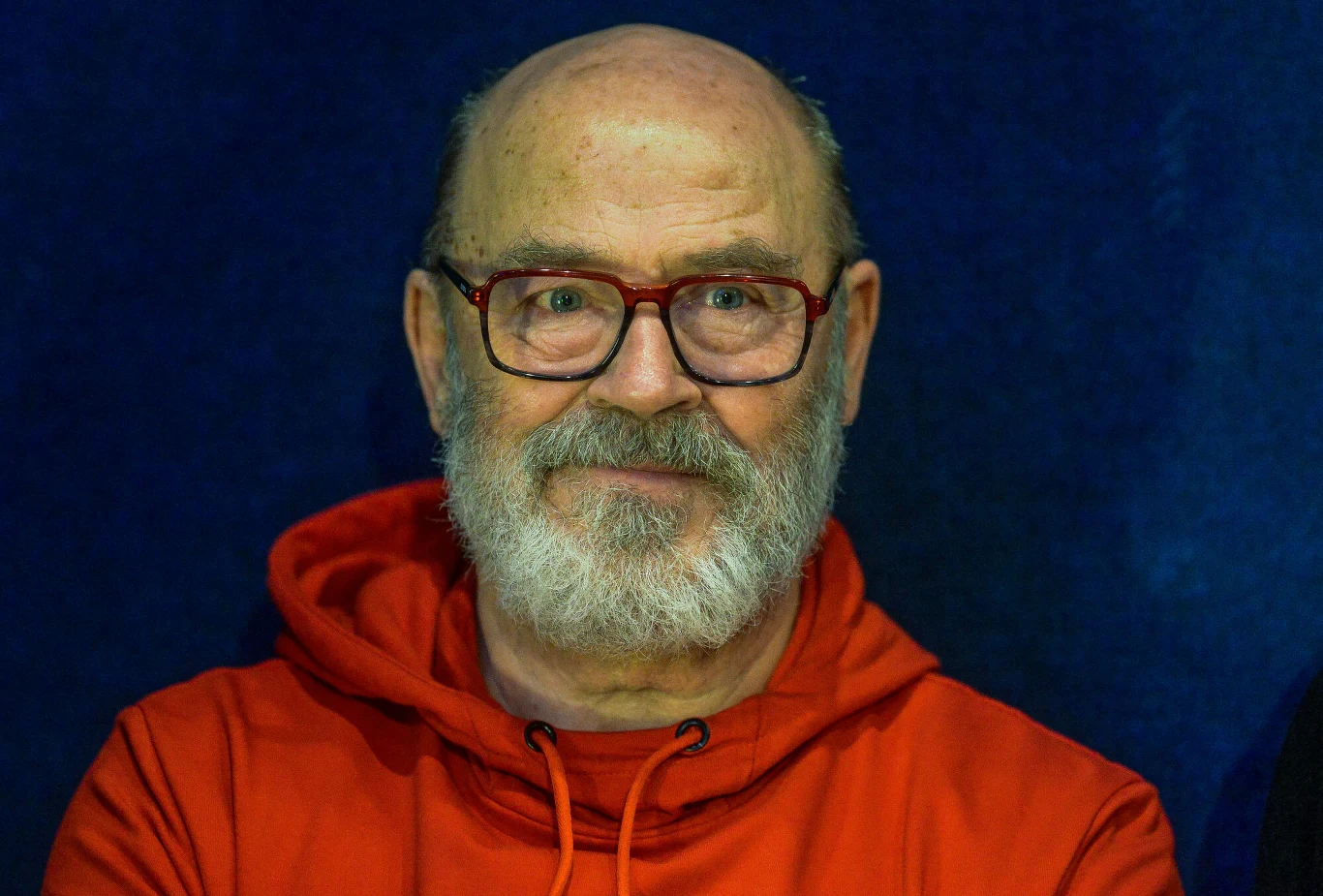 Jan Jakub Kolski Starszy mężczyzna z brodą oraz okularami w czerwonych oprawkach, ubrany w czerwoną bluzę z kapturem, na ciemnoniebieskim tle.