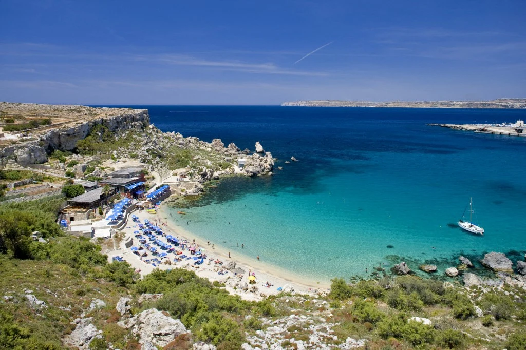 W kwietniu można wybrać się na wakacje na Maltę. Ceny all inclusive zaczynają się od 2 642 zł. Szeroka, piaszczysta plaża z leżakami oraz parasolami przyturkusowej wodzie, otoczona skałami i zielenią, z zabudowaniami przy samym brzegu i zacumowaną żaglówką na wodzie.