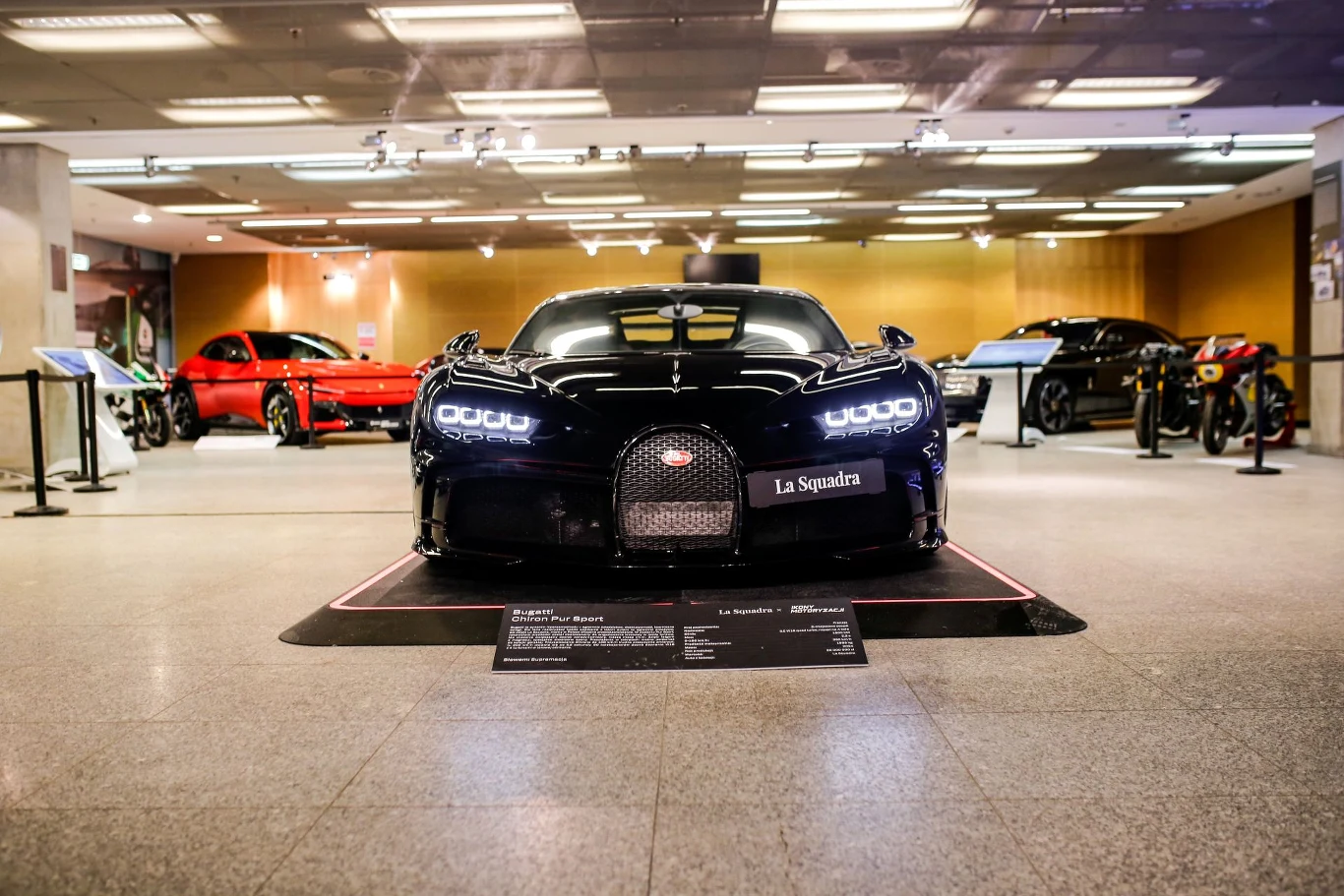 Luksusowy, czarny samochód sportowy Bugatti ustawiony na wystawie w dobrze oświetlonym salonie, w tle inne samochody sportowe, widoczne informacyjne tablice ekspozycyjne oraz nowoczesne wnętrze.