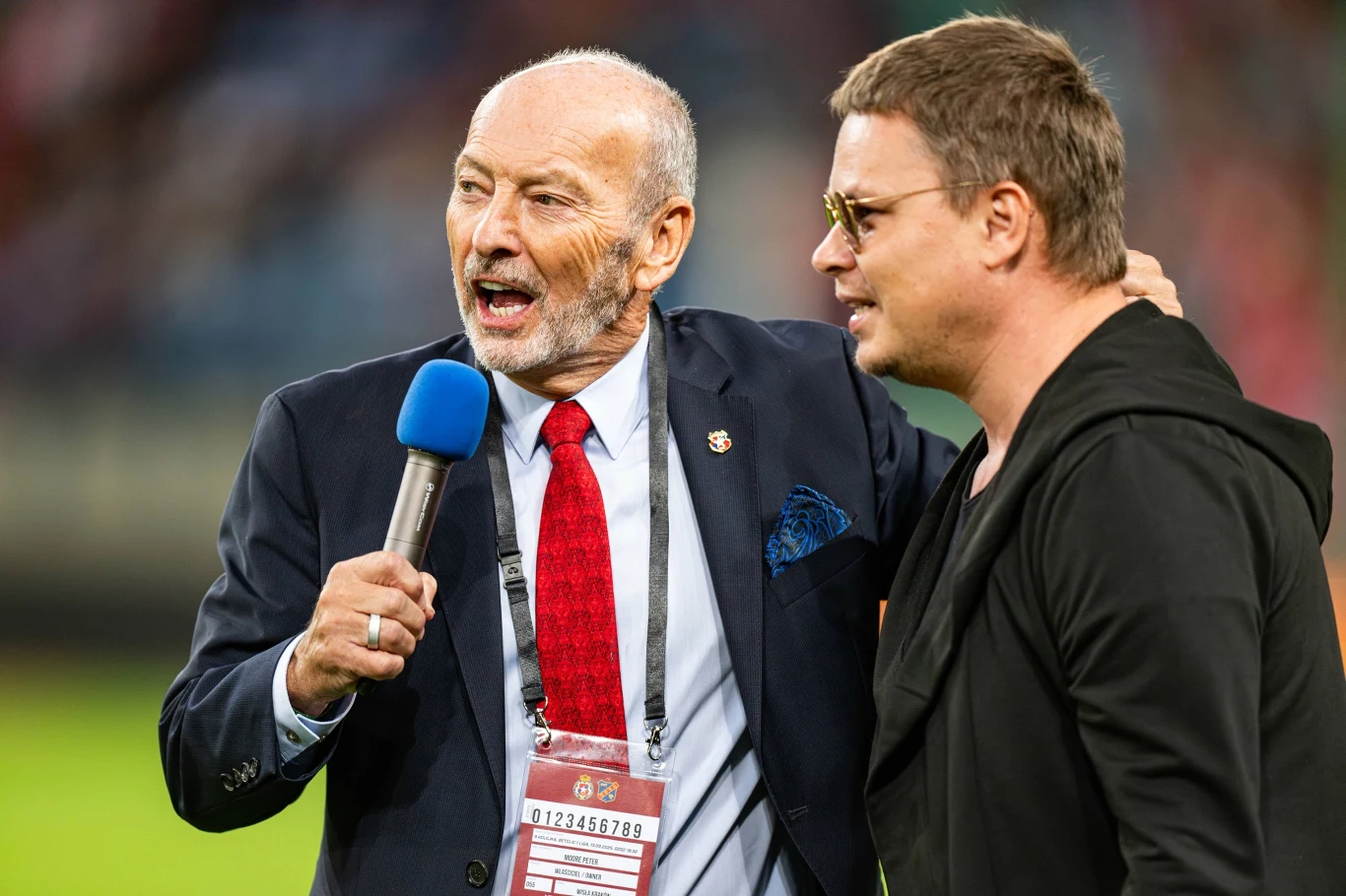 Dwóch mężczyzn w centrum stadionu, jeden w garniturze z mikrofonem, drugi w okularach i ciemnej bluzie, stoją blisko siebie, w tle widać rozmazane trybuny pełne kibiców.