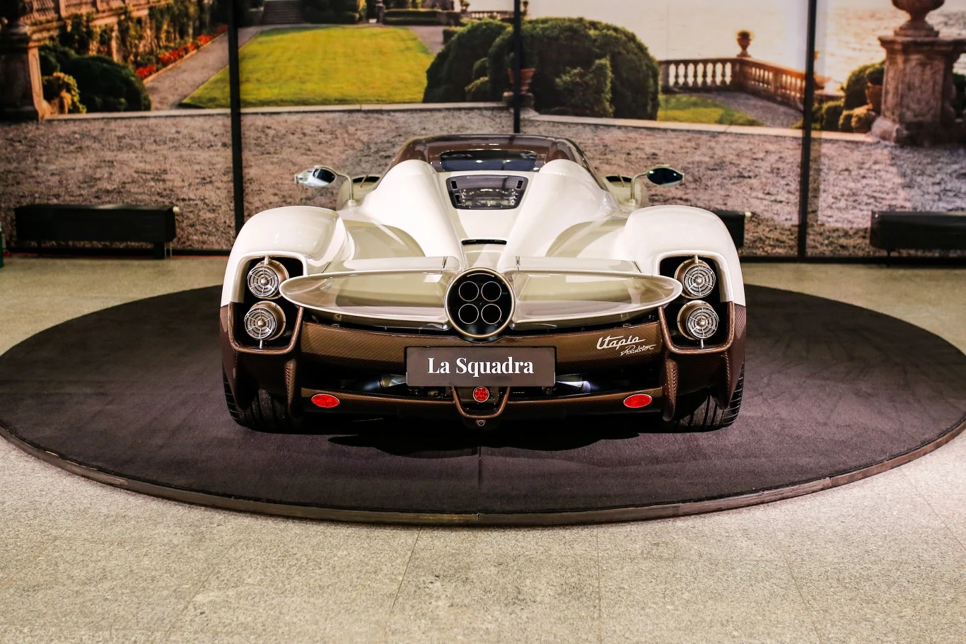 Sportowy samochód Pagani Utopia w białym kolorze ustawiony na ciemnym okrągłym dywanie, widok od tyłu, charakterystyczny układ wydechu z centralnie umieszczonymi rurami oraz LED-owe tylne światła, tablica z napisem La Squadra.