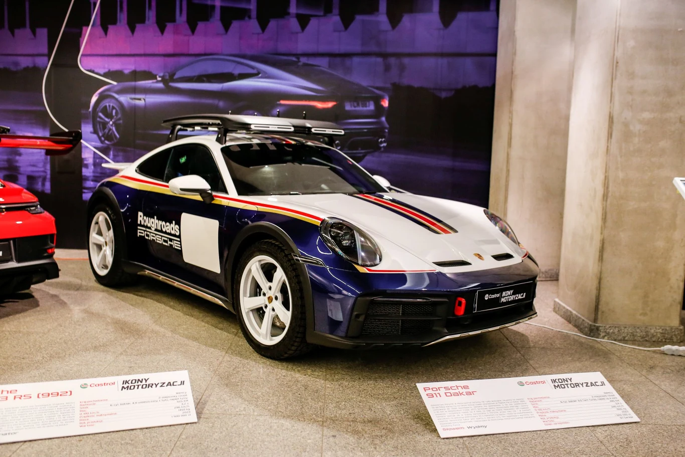 Samochód sportowy Porsche z napisem Rallyroads Performance, zaprezentowany na ekspozycji, stoi na tle podświetlonej planszy z sylwetką auta. Model posiada wyraziste barwy i sportowe pasy na masce.