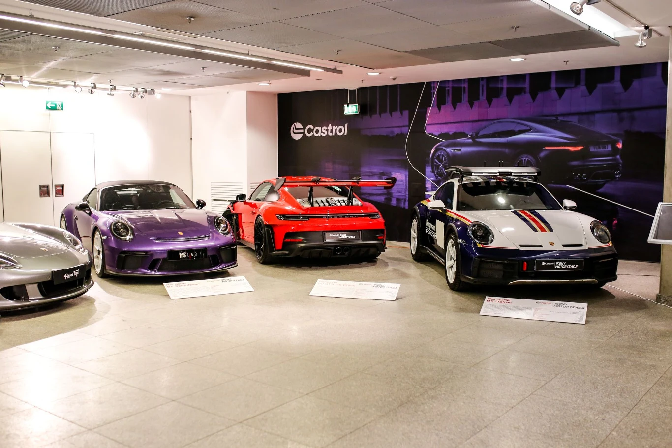 Trzy sportowe samochody Porsche w różnych wersjach kolorystycznych, ustawione obok siebie w jasnej galerii wystawowej. W tle baner z logo Castrol i grafika przedstawiająca sylwetkę samochodu.