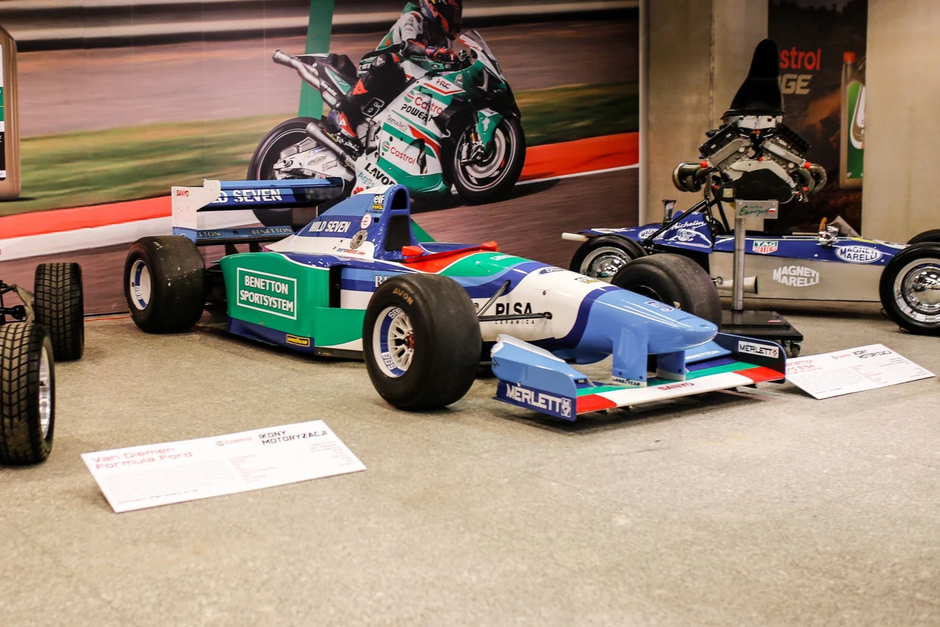 Bolid wyścigowy Formuły 1 w kolorach niebiesko-zielono-białych z nazwami sponsorów na karoserii ustawiony na ekspozycji wewnętrznej. W tle motocykl sportowy na plakacie oraz inne pojazdy eksponowane.