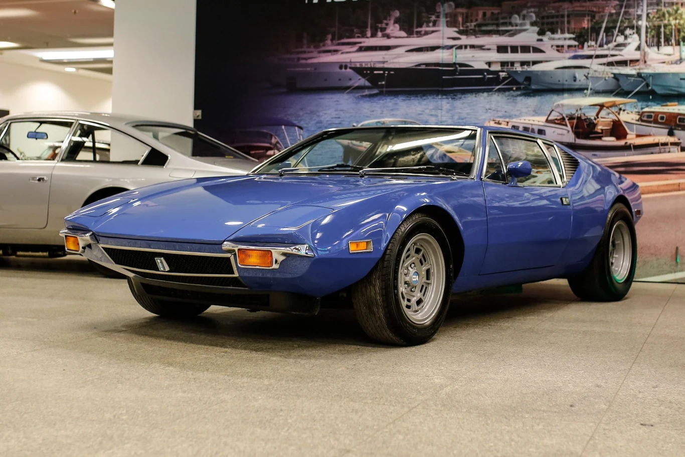 Klasyczny, niebieski samochód sportowy De Tomaso Pantera z lat 70., stojący w przestronnym pomieszczeniu z innymi samochodami i dużym zdjęciem jachtów w tle.