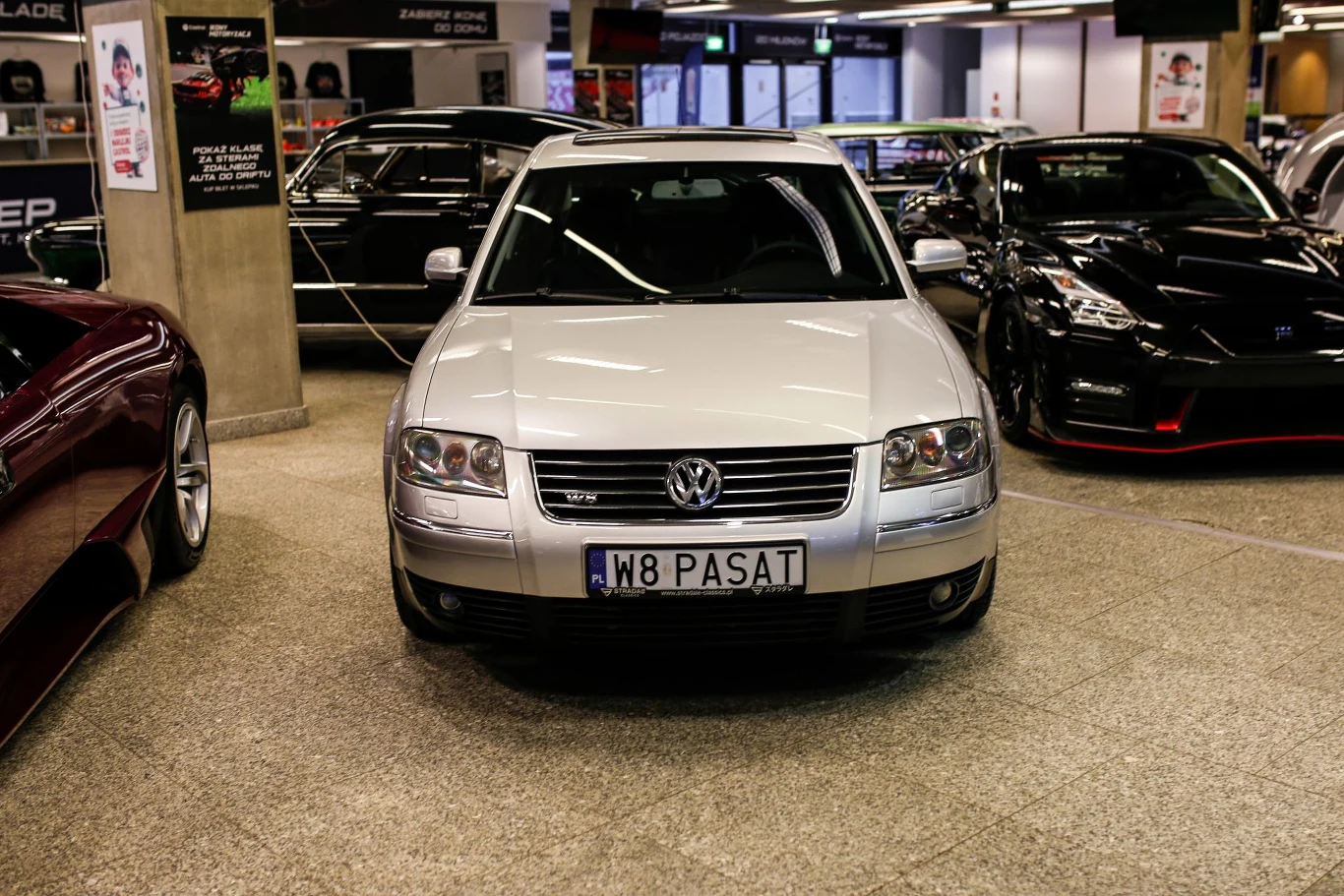 Srebrny Volkswagen Passat ustawiony przodem na wystawie motoryzacyjnej, otoczony innymi samochodami, z widoczną spersonalizowaną tablicą rejestracyjną W8 PASAT.