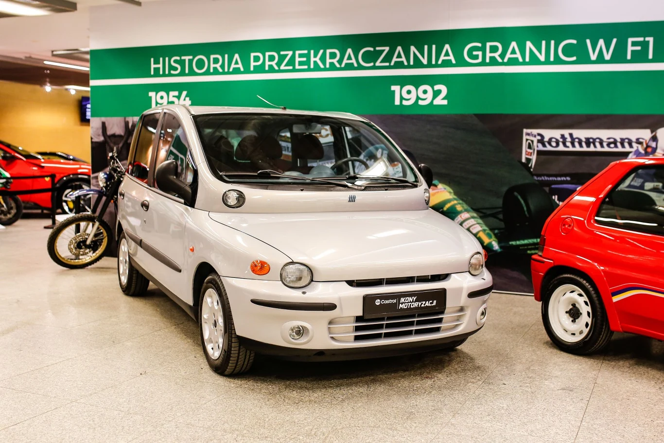 Srebrny samochód Fiat Multipla stoi w muzeum motoryzacji, otoczony innymi pojazdami, w tle zielony baner z napisem Historia przekraczania granic w F1 oraz datami 1954 i 1992.