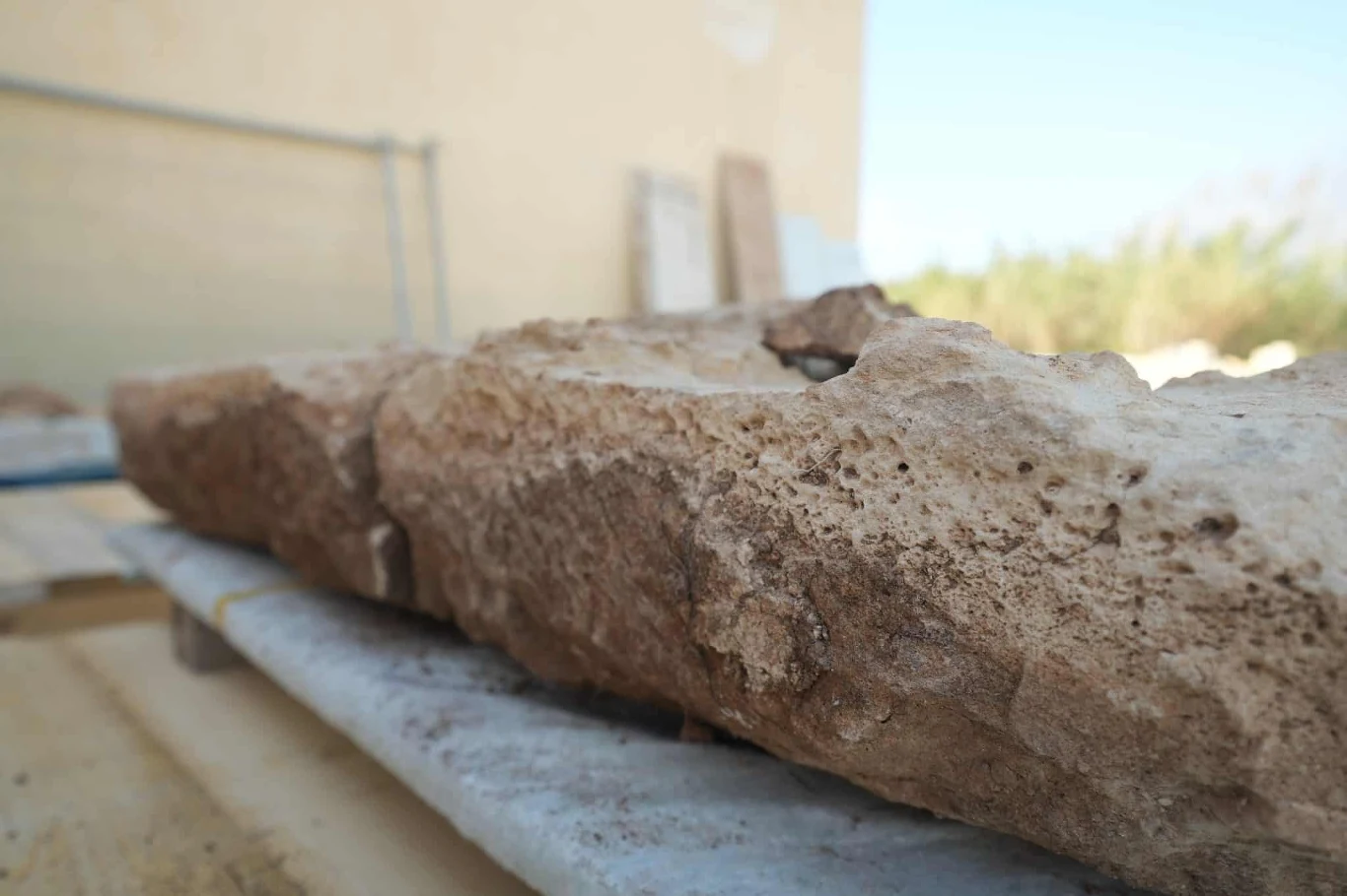 Fragment dużego, nieregularnego bloku kamiennego leżącego na marmurowej płycie, w tle rozmazana ściana oraz elementy placu budowy.