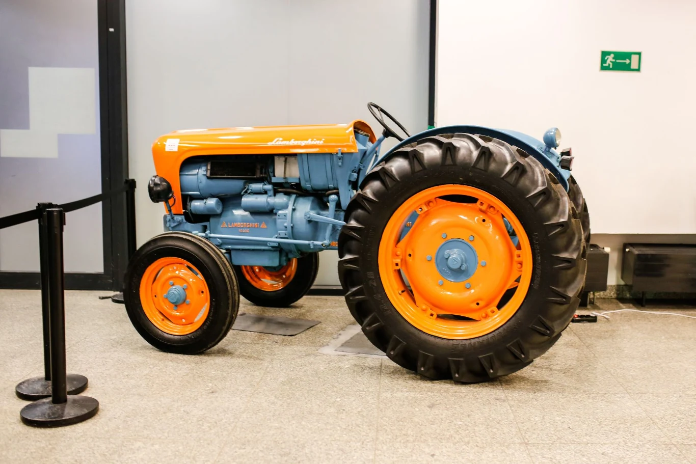 Zabytkowy traktor Lamborghini w kolorach niebieskim i pomarańczowym wystawiony w pomieszczeniu, stojący na jasnej podłodze, obok barierek ochronnych i tabliczki z napisem.