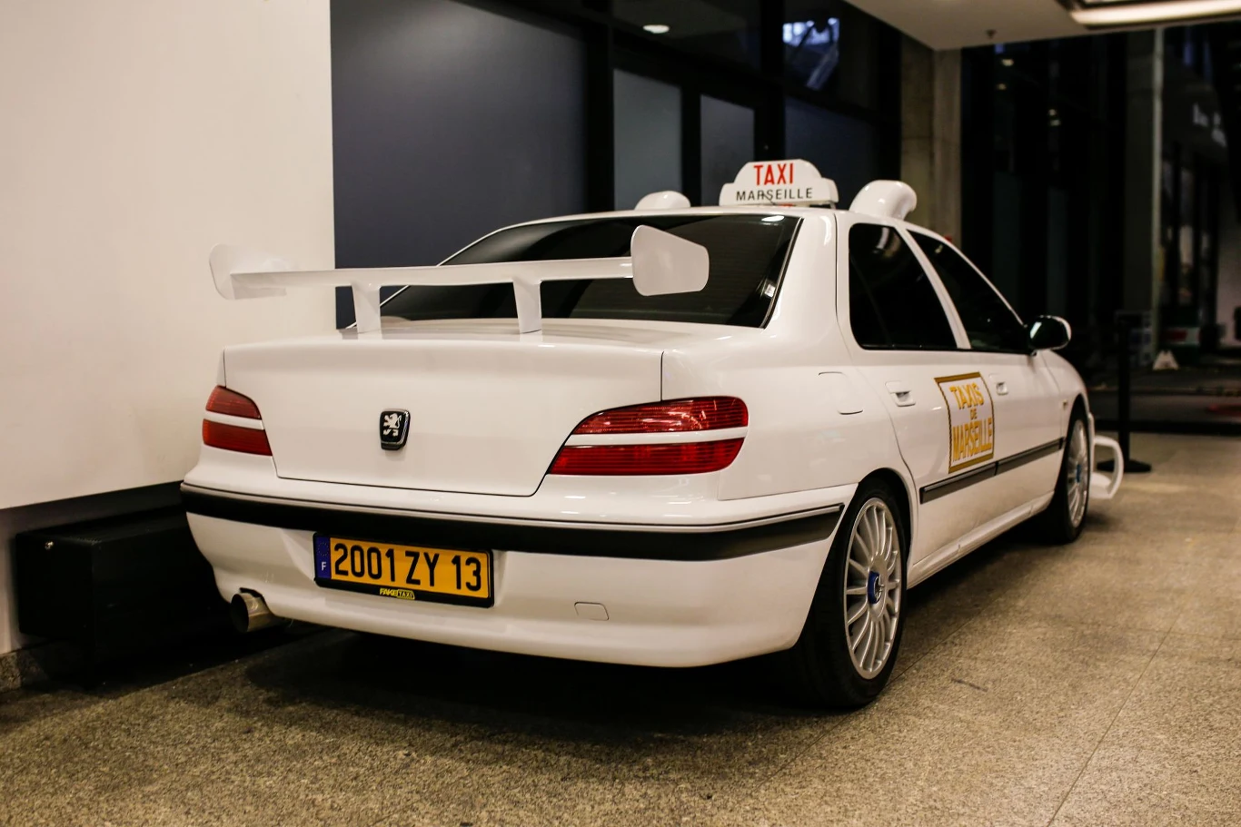 Białe sportowe auto z charakterystycznym tylnym spojlerem, żółtą tablicą rejestracyjną i oznaczeniem taxi na dachu, ustawione wewnątrz budynku, oświetlone sztucznym światłem.
