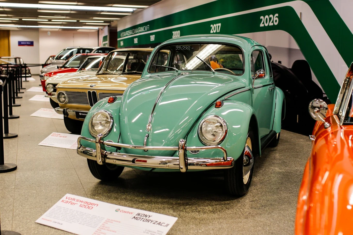 Kultowy, jasnozielony Volkswagen Garbus eksponowany na wystawie klasycznych samochodów, ustawiony obok innych zabytkowych aut w dobrze oświetlonej hali wystawienniczej. W tle na ścianie widoczne są napisy z datami, nawiązujące do historii motoryzacji.