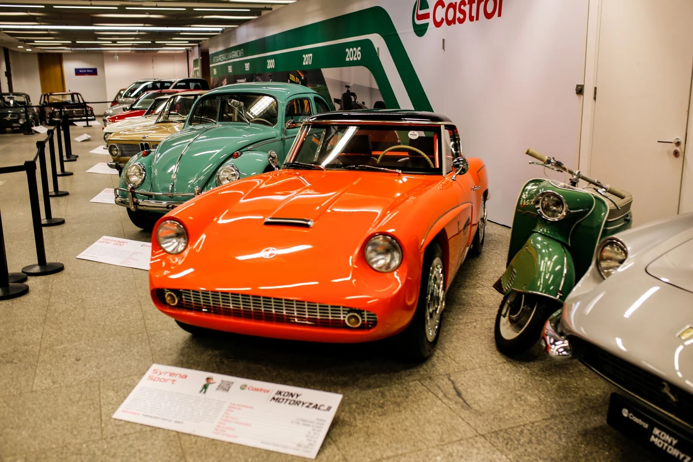 Wyeksponowany pomarańczowy samochód sportowy wśród innych klasycznych pojazdów na wystawie motoryzacyjnej, obok niego znajduje się zielony skuter. W tle widoczne są inne samochody oraz ściana z logo Castrol.