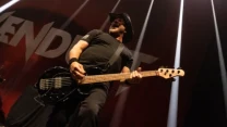 Muzyk ubrany na czarno grający na gitarze basowej na scenie podczas koncertu, dynamiczne światła sceniczne oraz widoczne logo zespołu w tle.
