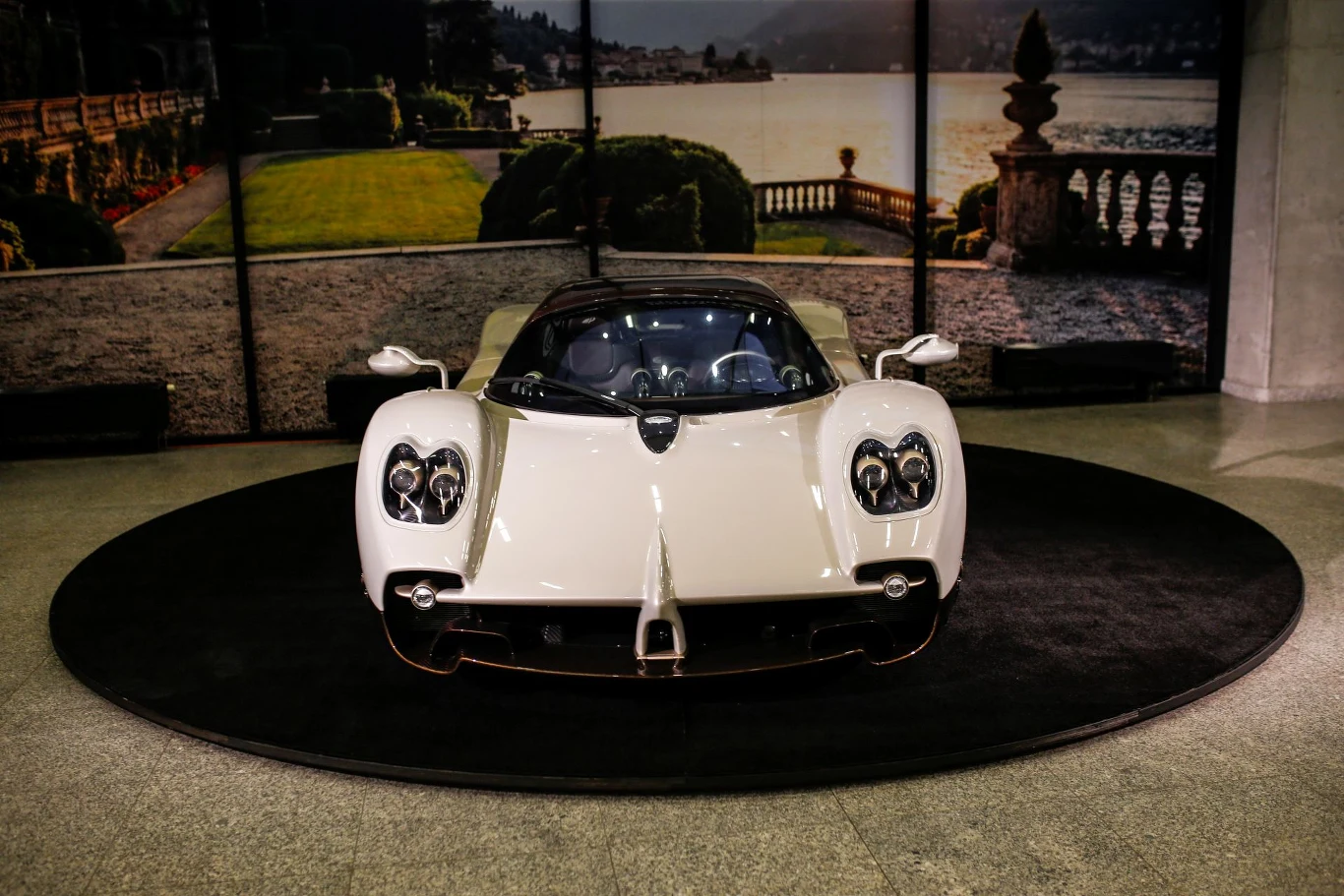 Białe ekskluzywne auto sportowe ustawione centralnie na czarnej okrągłej platformie, w tle panorama jeziora i ogrodu widoczna na dekoracyjnej ściance.