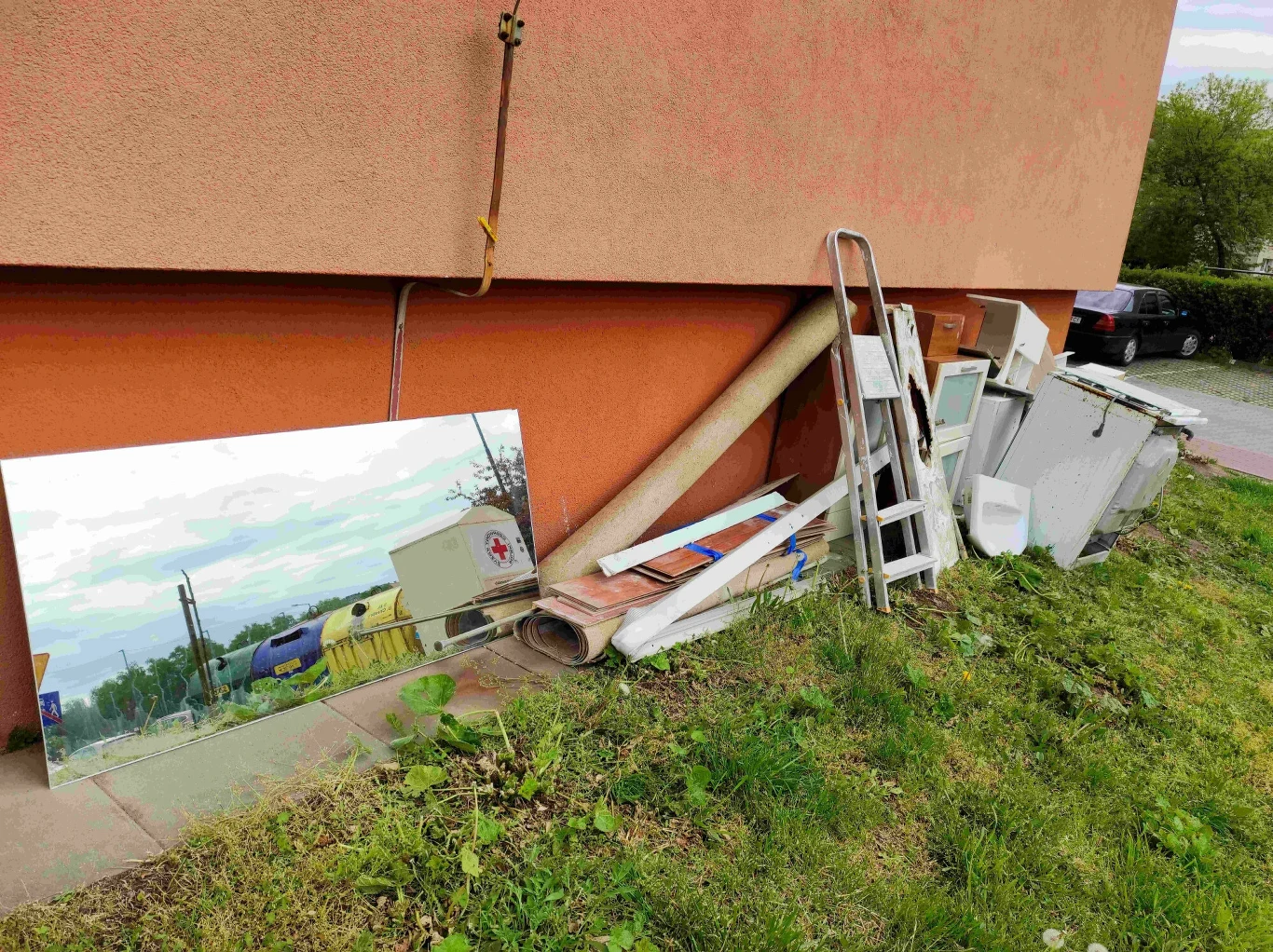 Sterta odpadów wielkogabarytowych pod ścianą budynku, obejmująca lustro, wykładzinę, drabinę, stare meble i inne przedmioty codziennego użytku, częściowo przysłonięta przez trawnik i chodnik.