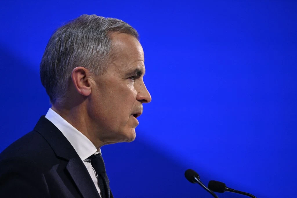 Premier Kanady Mark Carney podczas wystąpienia na Światowym Forum Ekonomicznym w Davos Mężczyzna w garniturze przemawia na tle jednolitej, niebieskiej sceny. Widoczny mikrofon sugeruje wystąpienie publiczne, światło skierowane na jego twarz podkreśla powagę sytuacji.