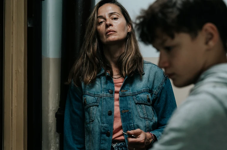 Kobieta w dżinsowej kurtce o poważnym wyrazie twarzy stoi przy ścianie, na pierwszym planie nieostro widoczny młody chłopak z zamyślonym spojrzeniem.
