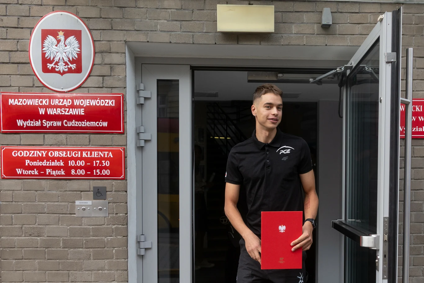 Młody mężczyzna w czarnym stroju sportowym stoi przed wejściem do Mazowieckiego Urzędu Wojewódzkiego w Warszawie, trzymając w rękach czerwoną teczkę z godłem Polski. Na ścianie widoczne są tablice informacyjne z nazwą urzędu oraz godzinami obsługi klie...