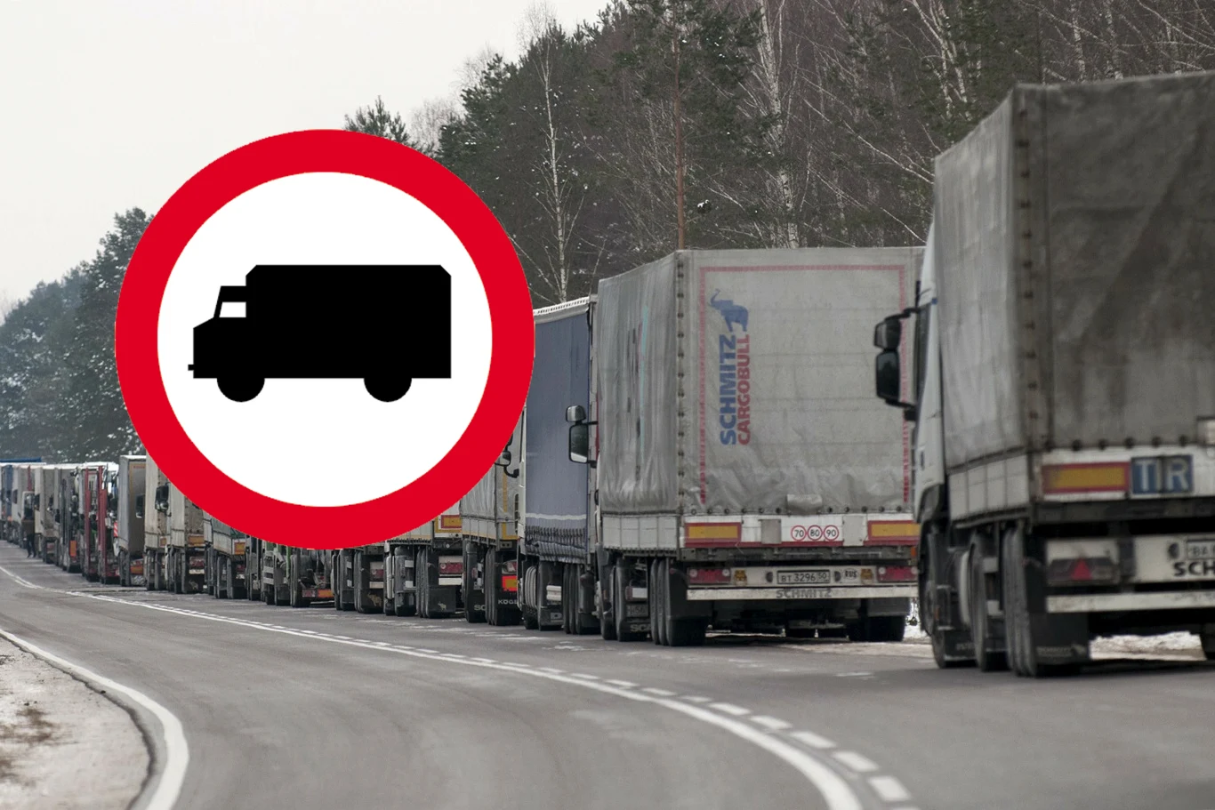 Nowe ograniczenia dla ciężarówek. Uwaga w regionie Igrzysk Olimpijskich Długa kolejka ciężarówek stojących na drodze z zakrętem, zimowy krajobraz, pobocze przykryte śniegiem, przy drodze stoi znak zakazu wjazdu dla pojazdów ciężarowych, w tle drzewa bez liści.