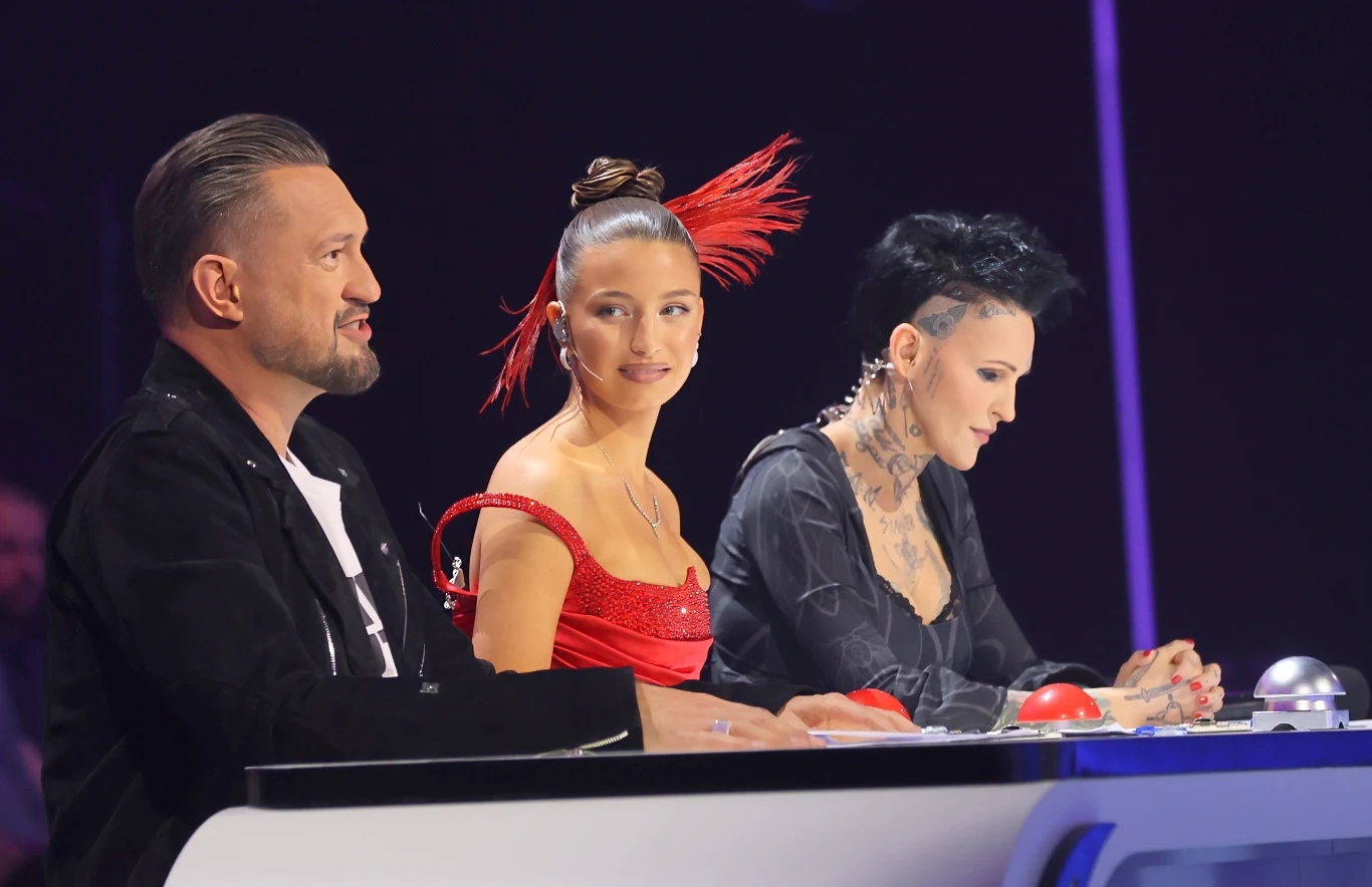 "Mam Talent!" powraca! Trójka jurorów siedzi przy stole, przed nimi kolorowe przyciski, każdy skierowany w stronę sceny. Kobieta pośrodku ma elegancką czerwoną suknię i ozdobę we włosach, pozostali jurorzy ubrani są na ciemno, z widocznymi tatuażami i charakterystycznymi fry...