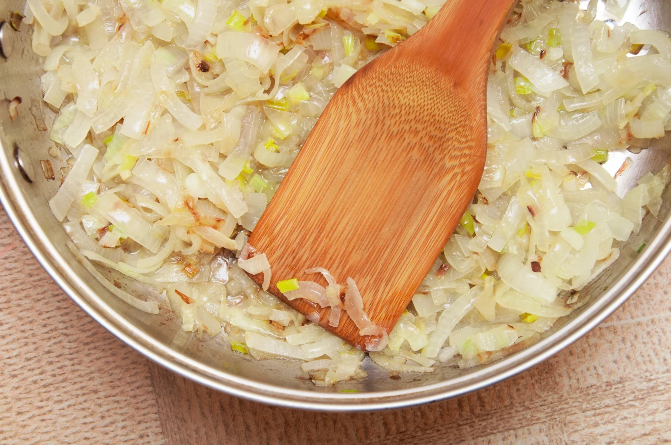 Smażona cebula na patelni mieszana drewnianą łopatką, delikatnie zarumienione kawałki cebuli i lekko złocisty kolor świadczący o procesie podsmażania.