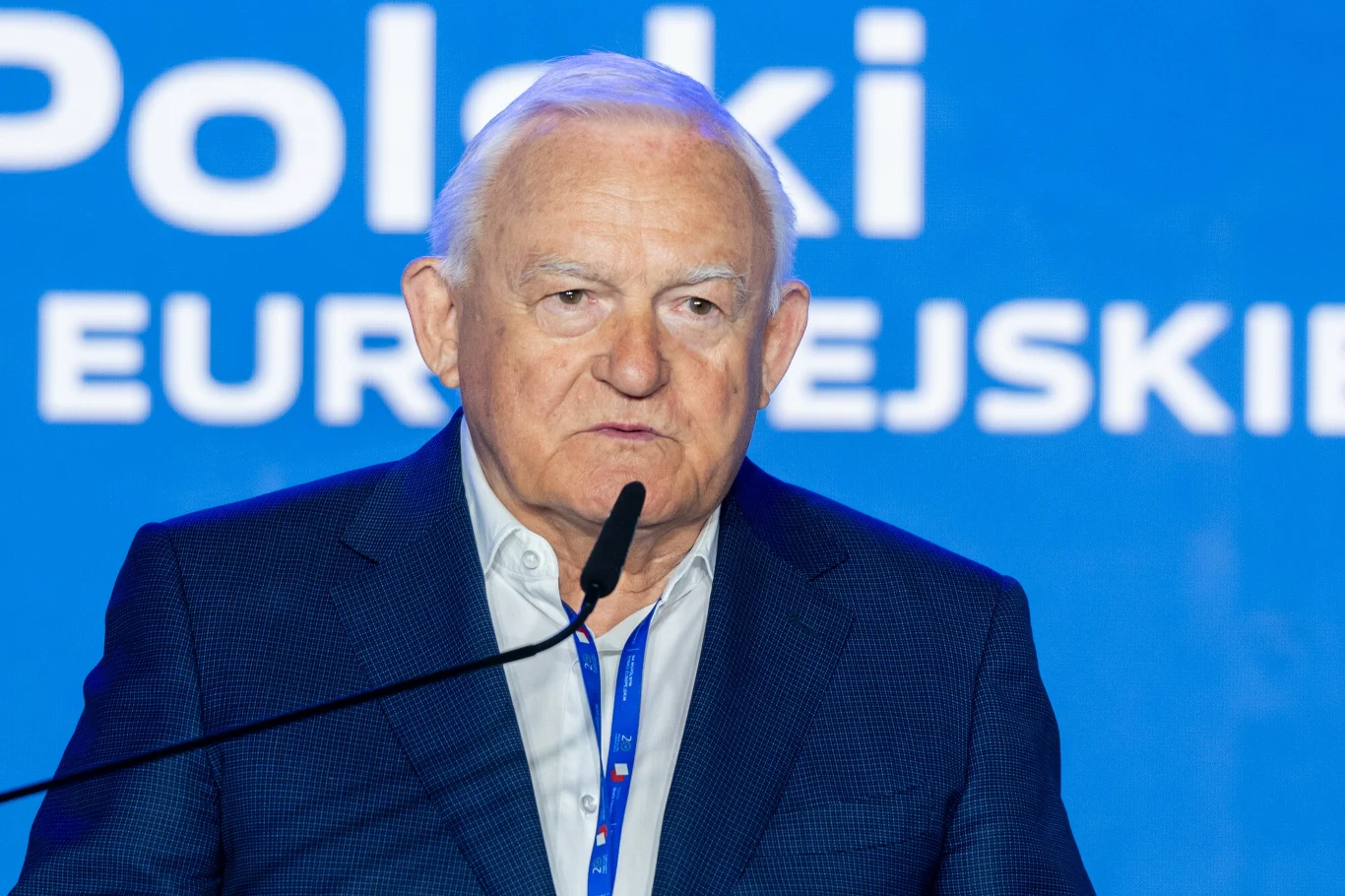 Starszy mężczyzna w garniturze przemawia do mikrofonu na tle niebieskiej ścianki z napisem.