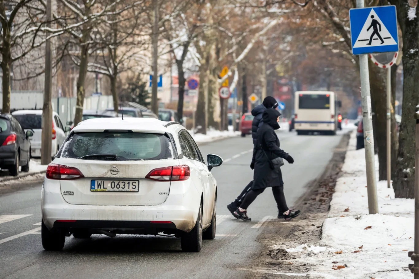 Czy można ruszyć, kiedy pieszy znajduje się jeszcze na przejściu? Osoba ubrana w ciemną kurtkę i czapkę przechodzi przez przejście dla pieszych na zaśnieżonej ulicy, obok przejeżdża biały samochód, w tle widoczne są znaki drogowe i autobus.