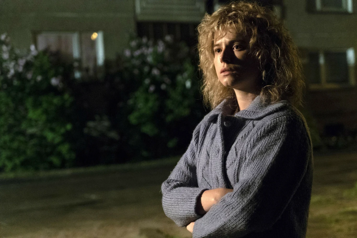 Jessie Buckley w "Czarnobylu" Kobieta z jasnymi, falowanymi włosami stoi na tle rozmytej ulicy oraz zieleni, ubrana w grubą szarą kurtkę, z założonymi rękami i poważnym wyrazem twarzy, oświetlona miękkim światłem.