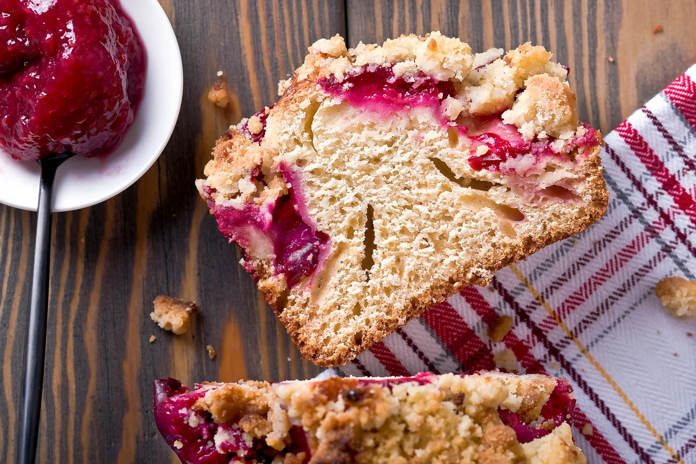 Kawałek drożdżowego ciasta z widocznym przekrojem i dodatkiem owoców, posypany chrupiącą kruszonką, obok na białej miseczce leży porcja dżemu, a całość znajduje się na drewnianym blacie z fragmentem lnianej serwetki w czerwone pasy.