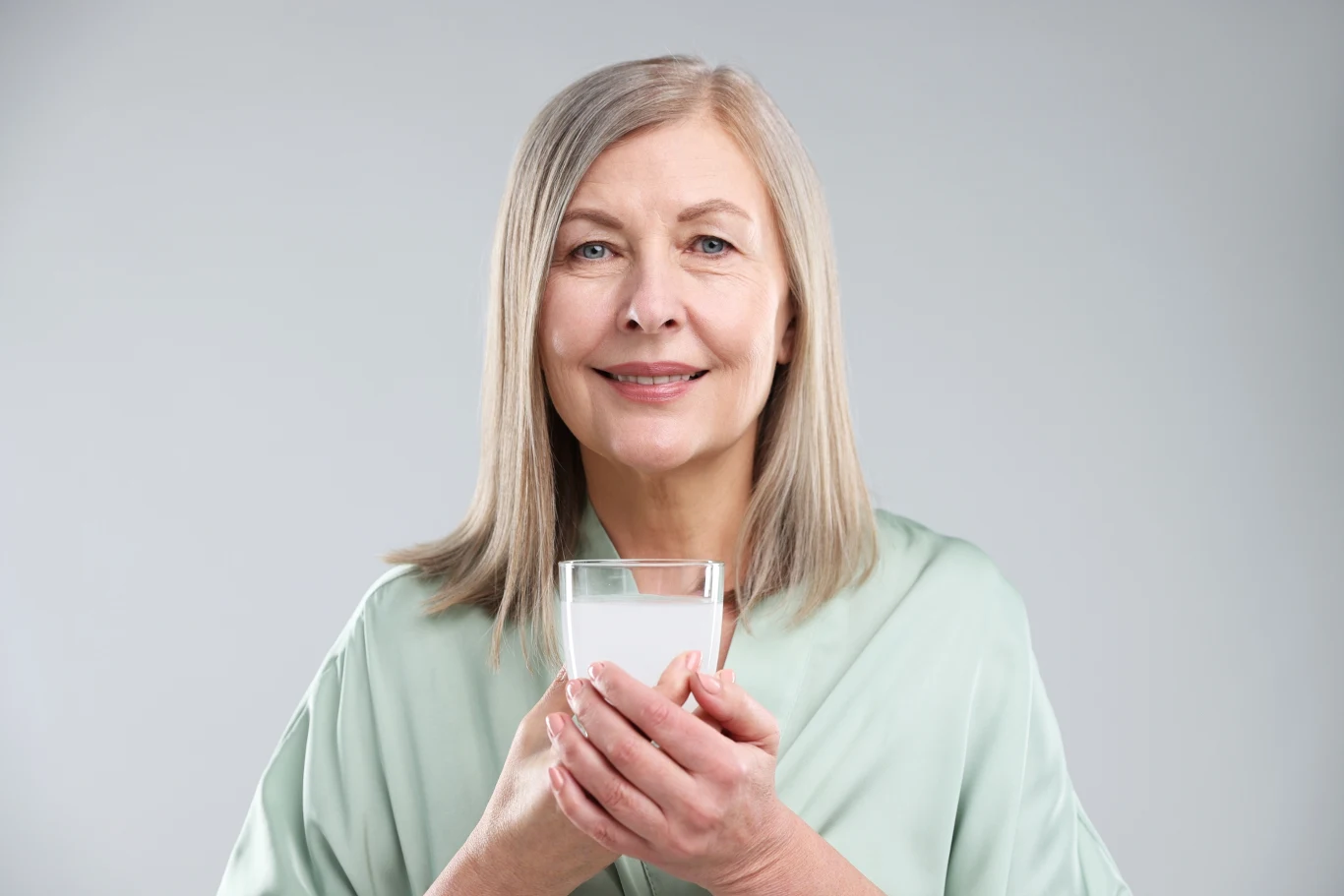 Starsza kobieta o siwych włosach w jasnym ubraniu trzyma w dłoniach szklankę mleka i uśmiecha się delikatnie do obiektywu na jasnym tle.
