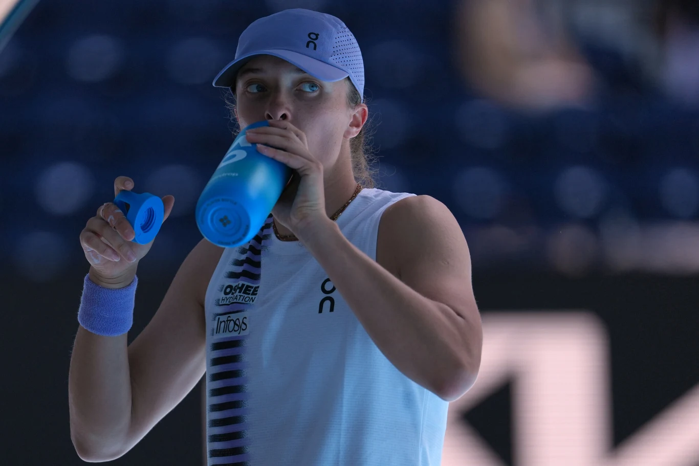 Tenisistka w sportowej koszulce i czapce na głowie, pijąca wodę z niebieskiego bidonu podczas przerwy w meczu tenisowym na korcie.