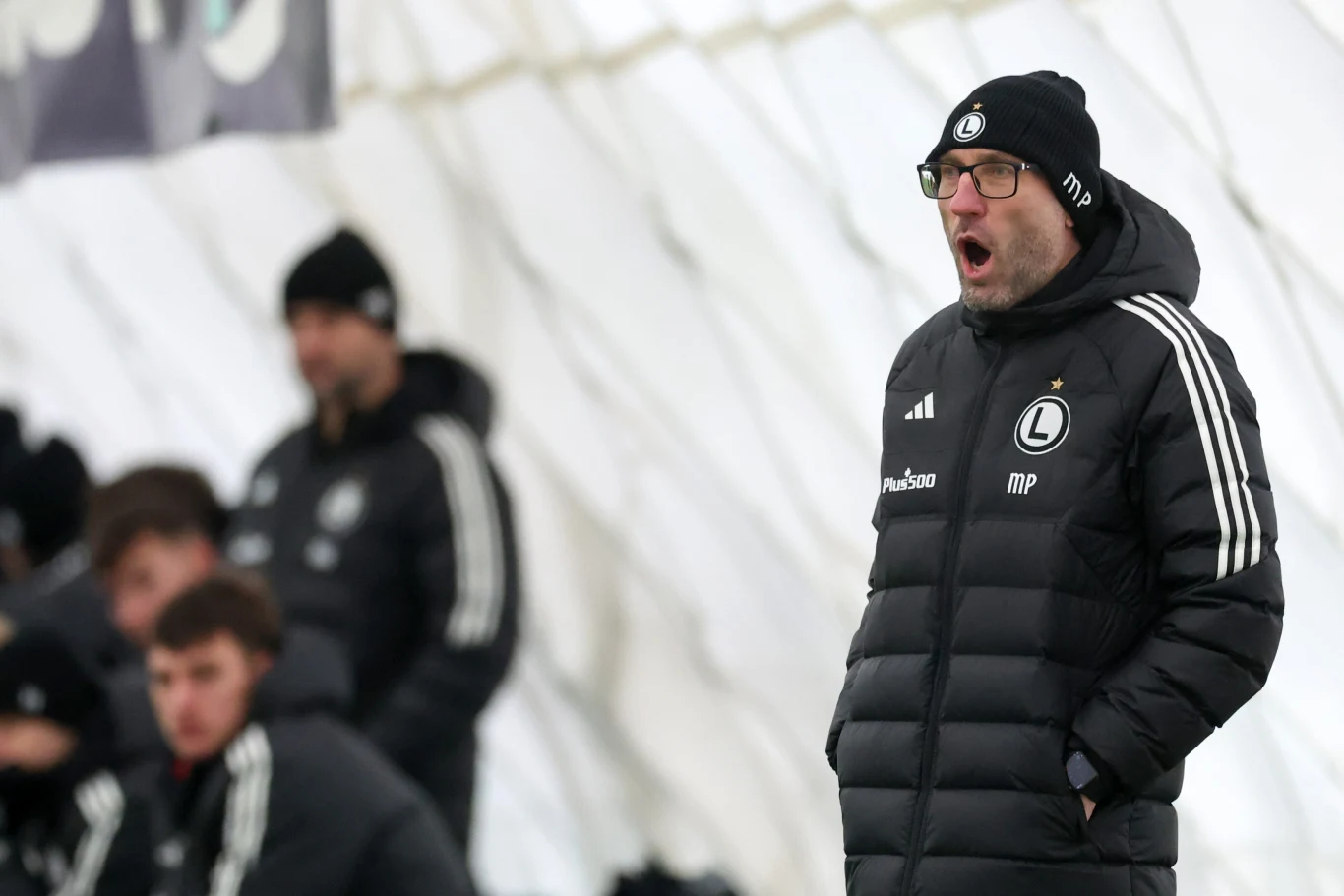 Marek Papszun Trener ubrany w czarną kurtkę i czapkę zimową z logo klubu Legia Warszawa stoi z otwartymi ustami, prawdopodobnie krzycząc lub udzielając wskazówek, na tle innych osób siedzących na ławce.