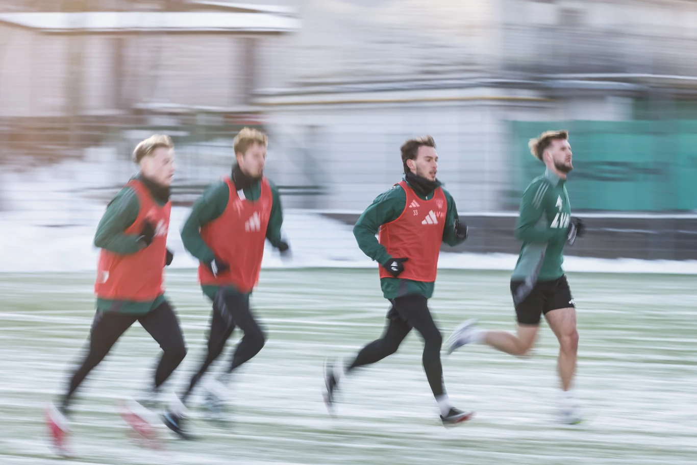 Piłkarze Ekstraklasy wrócili z cieplejszych krajów, gdzie jak co rok bawili na obozach przygotowawczych. Zderzenie z pogodowymi realiami jest bolesne Czterech młodych mężczyzn biegnie po boisku sportowym, ubrani w zielone stroje oraz czerwone kamizelki treningowe, w tle widać rozmyte elementy zabudowy i zimową aurę.
