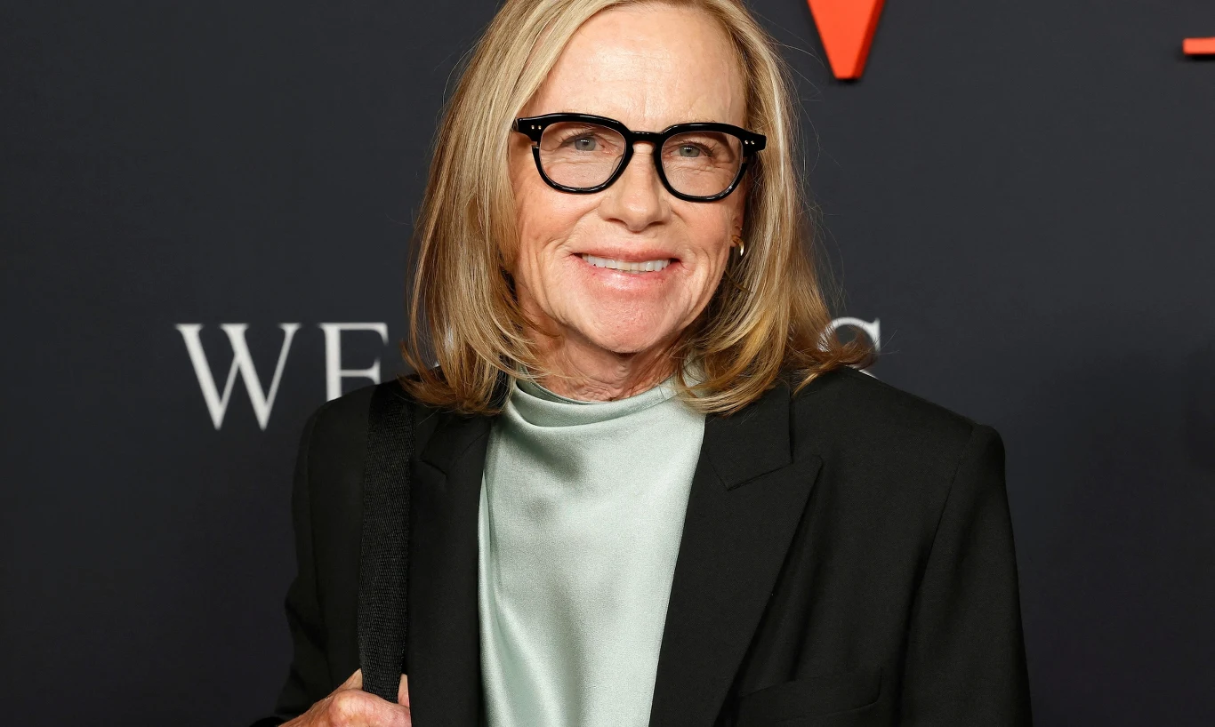 Amy Madigan Uśmiechnięta kobieta w okularach i eleganckiej marynarce na tle ciemnej ścianki z dużymi literami w tle.