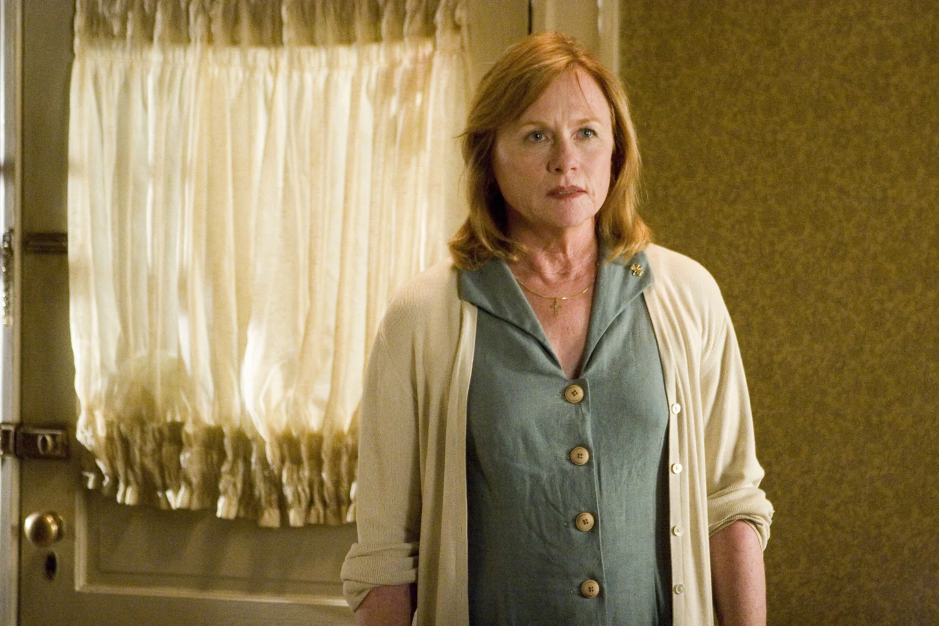 Amy Madigan w filmie "Gdzie jesteś, Amando?" Kobieta o rudych włosach w niebieskiej sukience i kremowym swetrze stoi przy drzwi z firanką, ma poważny wyraz twarzy, stoi w domowym wnętrzu o ciepłych kolorach ścian.